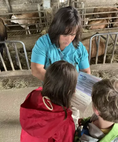 Mujer mostrando un documento a dos niños, y un grupo de vacas en la parte de atrás